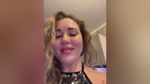 Media: Video of a smiling, light-skinned woman with wavy, shoulder-length blonde hair, wearing a black sequined top. She has subtle makeup, with pink lips and subtle eyeshadow. Background is a blurred beige wall.