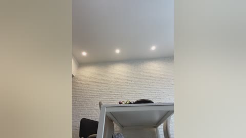A video of a modern dining room with a white brick wall, four recessed ceiling lights, a light gray dining table, and a black chair with a cushion. The room is bright and minimalist.