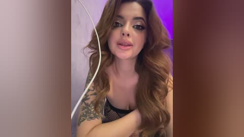 Media: Video of a woman with long, wavy brown hair, wearing a black lace top, showcasing tattoos on her left arm. She's standing against a blurred, gradient purple and brown background.