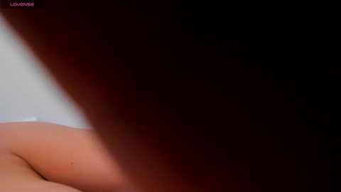A close-up video of a nude woman's back and buttocks, with a shadowy, dark background, showcasing smooth, pale skin and a hint of a tattoo. The image has a soft, sensual atmosphere.