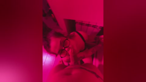 Media: A dimly lit video captures a woman with short blonde hair, wearing glasses and black lingerie, lying on a bed under a red glow. Her face is partially visible, with a relaxed expression. The background includes a patterned bedspread and a nightstand.