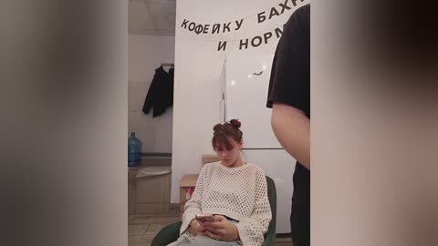 Media: Video of a young woman with dark hair in a bun, wearing a white crochet sweater, sitting in a green chair, texting, in a dimly lit, cluttered room with a Russian-language banner and a water jug.