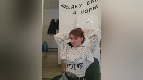 Media: Video of a young woman with light skin and brown hair, wearing a white crocheted sweater and jeans, sitting in a room with a white cabinet and black coat hanging on a hook.