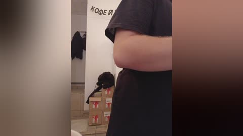 Media: Video of a person in a black shirt, standing with arms crossed, partially visible. Background includes a wall with a sign and a pile of cardboard boxes.