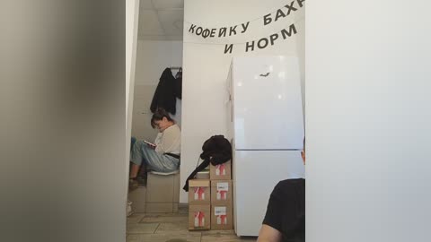 Media: A video of a small, sparsely furnished room with two people sitting on a floor, reading books. The room has white walls and a fridge with Cyrillic text.