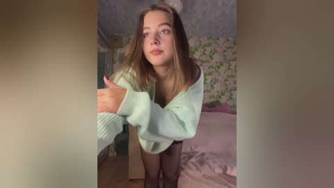 A video of a young Caucasian woman with straight, shoulder-length brown hair, wearing a light green knitted sweater and black stockings, leaning forward in a bedroom with floral wallpaper and pink bedding.