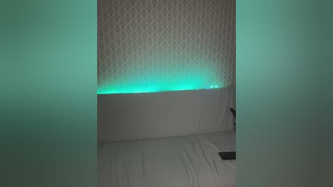 A video of a dimly lit bedroom with white bedding and a textured wallpaper backdrop. A soft green LED strip lights the headboard area. The room appears minimalistic and serene.