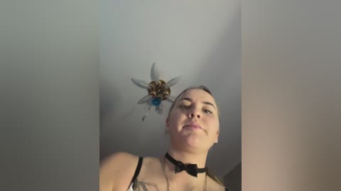 Media: A video of a woman with fair skin, short hair, and a black choker, wearing a black bra and bow tie. She's looking up at a decorative fairy hanging from the ceiling. The background is plain white.