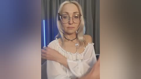 Media: Video of a middle-aged Caucasian woman with shoulder-length platinum blonde hair, wearing glasses, a white off-shoulder top, and a choker necklace. She holds a blue light saber, set against a blurred gray curtain background.