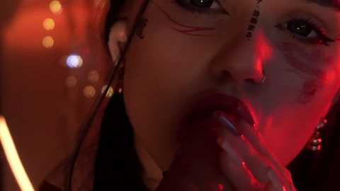 Media: Close-up video of a woman with a dark complexion, wearing red lipstick, red eye makeup, and a nose ring, illuminated by red light, with blurred city lights in the background.