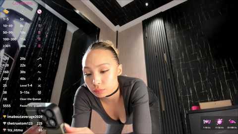 Video of an Asian woman with light skin and brown hair, wearing a black choker and top, focused on a phone screen with social media stats in a dimly lit room.
