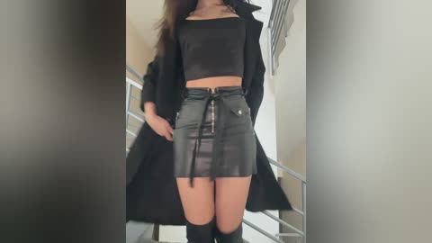 Media: Video of a woman in a black leather crop top and high-waisted leather skirt, wearing a long black coat, standing in a modern, minimalist hallway with metal railings.