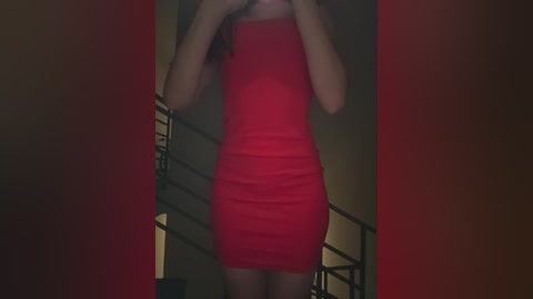 Media: Video of a light-skinned woman with long, wavy hair, wearing a tight, bright red, sleeveless mini dress, standing in a dimly lit, narrow hallway with a metal railing on the left.