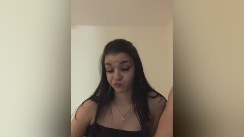 Video of a young woman with long black hair and fair skin, wearing a black strapless top, standing against a plain beige wall.