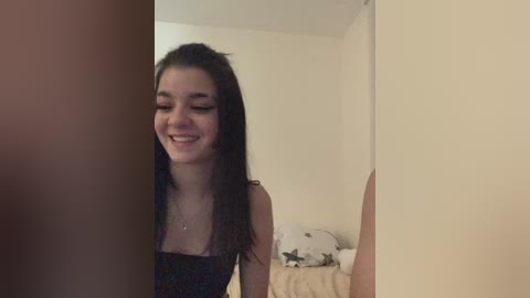 Video of a smiling young woman with long dark hair, wearing a black dress, standing in a dimly lit bedroom with beige walls and a messy bed.