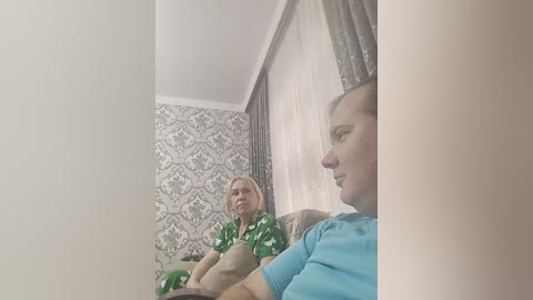 Media: Video of a woman with blonde hair in a green floral dress sitting on a sofa, looking at a man with brown hair in a blue shirt, both in a room with patterned wallpaper and sheer curtains.
