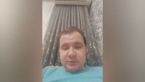 Media: Video of a middle-aged man with short brown hair, wearing a blue t-shirt, lying in a hospital bed with gray curtains and white bedding.