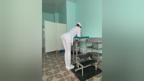 Media: Video of a female nurse in a hospital room, wearing white uniform and cap, bending over a patient on a hospital bed. The room has light green walls, a patterned floor, and medical equipment.