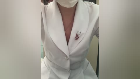 Media: Video of a woman in a white lab coat, wearing a medical mask, and a silver pin on the lapel, standing in a clinical setting with beige walls.