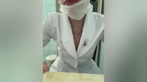 Media: Video of a person in a white lab coat, wearing a surgical mask and gloves, holding a clipboard, in a tiled bathroom with a toilet and cup.