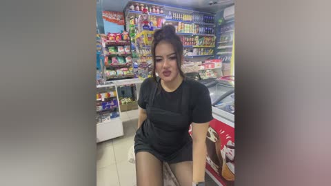 Media: Video of an Asian woman with dark hair, wearing a black short-sleeved top and shorts, sitting on a white counter in a brightly lit convenience store filled with colorful snacks and beverages.