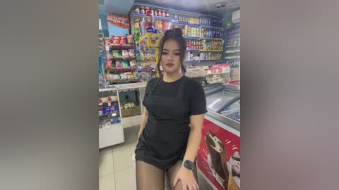 Media: Video of an Asian woman with medium build and dark hair styled in a bun, wearing a black dress, standing in a colorful convenience store.