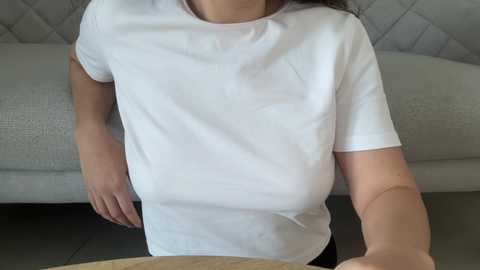 Video of a woman with light skin, wearing a white t-shirt, sitting on a grey, tufted sofa. She has a slim build and is looking down at a wooden table.