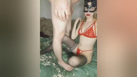 Media: Video of a man and woman in a bedroom. The man is nude, standing, with a light-skinned woman kneeling, wearing a red lace lingerie set, black stockings, and a black mask.