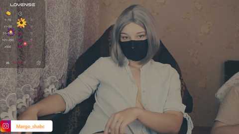 A video of a woman with shoulder-length silver hair, wearing a black face mask and a white blouse, sitting on a dark chair in a dimly lit room with patterned wallpaper.