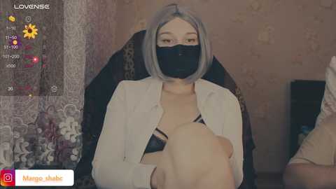A video of a woman with light skin and a short, gray bob haircut, wearing a black face mask, a white shirt, and a black bra, seated on a black chair in a room with floral wallpaper.