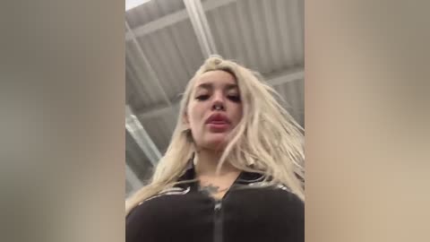 A video of a blonde woman with long hair and a black jacket, taken from a low angle, showing her large breasts and tattooed chest. The background features a metallic ceiling and industrial lighting.
