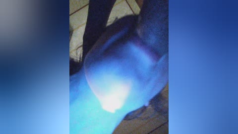 A video of a person, possibly a woman, with a dark complexion, standing in a dimly lit room with a blue light casting a glow on their face and torso. The background is a tiled floor.