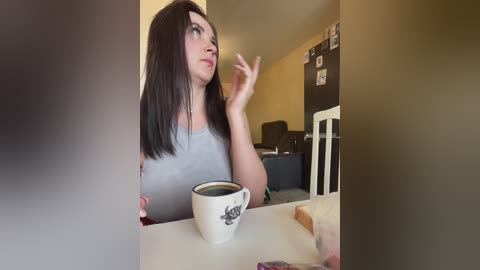 Media: Video of a woman with straight, shoulder-length black hair in a light grey top, holding a coffee cup, thinking in a cozy, yellow-painted room with framed photos and a blackboard.