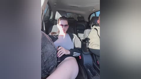 Media: Video of a light-skinned woman with straight, dark brown hair, wearing sunglasses and a grey top, sitting in a car.