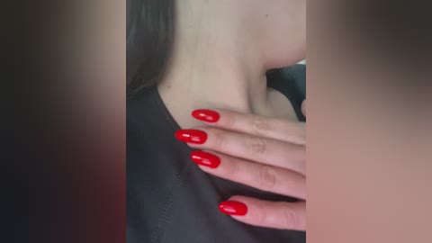 Media: Video of a woman with fair skin, wearing a black top, showcasing her long, glossy red fingernails. Her neck and collarbone are visible, suggesting a close-up shot. The background is blurred and dark.