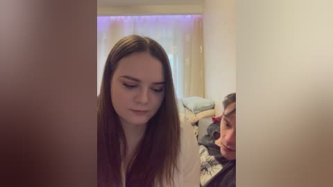 Video of a young woman with long, straight brown hair and fair skin, eyes closed, standing in a dimly lit bedroom. A man with short brown hair lies on a bed in the background.