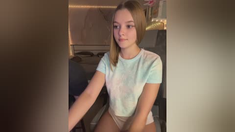 Media: Video of a young, light-skinned, blonde girl with straight hair, wearing a pastel tie-dye t-shirt and beige shorts, sitting indoors.