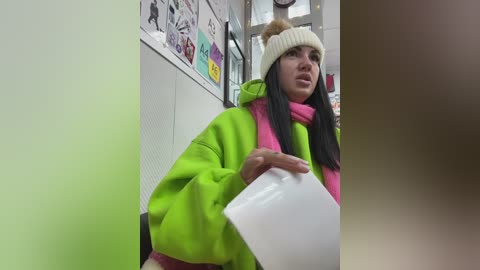 Video of a young woman with long black hair, wearing a bright green coat, beige hat, and pink scarf, holding a white mug in a dimly lit, cluttered indoor setting.