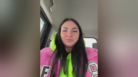 Video of a young woman with long, straight black hair, wearing a bright pink hoodie with a green hood, and sitting in a car with a blurred background.