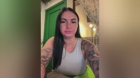 Video of a young woman with long black hair, wearing a white tank top, showcasing extensive tattoos on her arms. She stands in a hallway with green door and floral wallpaper, illuminated by warm lighting.