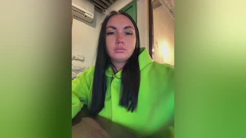 Video of a young woman with long black hair, wearing a bright green hoodie, standing indoors with blurred background, featuring a white wall, air conditioner, and a wooden door.