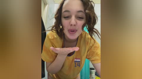 Media: Video of a young woman with wavy brown hair, wearing a yellow tie-dye shirt with a \"14\" on it, blowing a kiss while indoors.