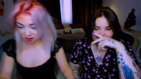 Video of two tattooed women in a dimly lit room, one with pink hair and red lipstick, the other with dark hair and a floral dress, both smiling.