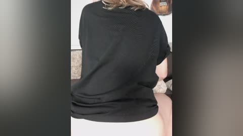 A video of a woman from behind, wearing a loose black shirt and white underwear, sitting on a bed in a dimly lit room.