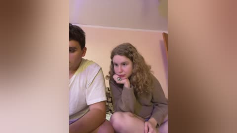 Media: Video of a young, fair-skinned girl with curly hair, in a beige sweater, leaning on a wall, with a boy in a white t-shirt, partially visible. Background features a patterned wall and a blurred ceiling light.