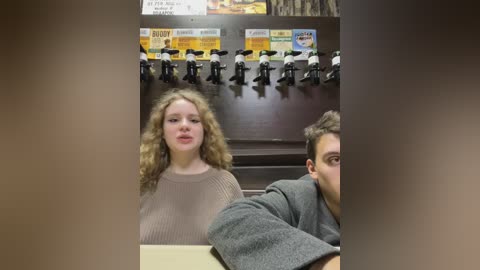 Media: Video of two people in a diner: a blonde woman with curly hair in a beige sweater, and a man in a gray sweater, seated at a booth. Behind them, a row of black-and-white photos and a menu board.