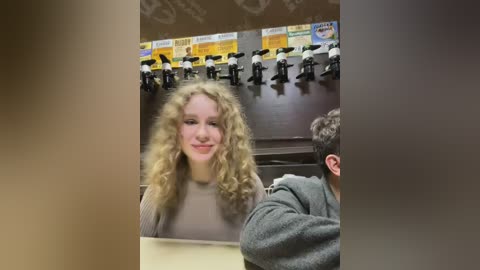 Media: Video of a young woman with curly blonde hair, smiling, wearing a beige sweater, seated at a desk with a gray background and a row of miniature figures in the background.