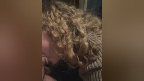 Media: A close-up video of a person with curly, shoulder-length blonde hair, partially obscured by a blurred background, wearing a beige knit sweater.