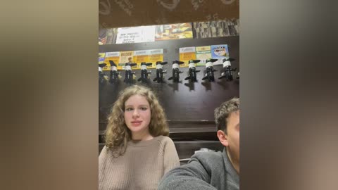 Media: Video of a young girl with curly hair and light skin in a beige sweater, seated indoors with a blurry man in the background.