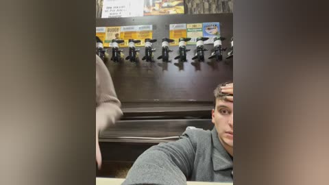 Media: A video shows a man with short dark hair, wearing a gray shirt, with his hand on his forehead, in a dark coffee shop. The background features a row of black coffee machines with colorful signs.